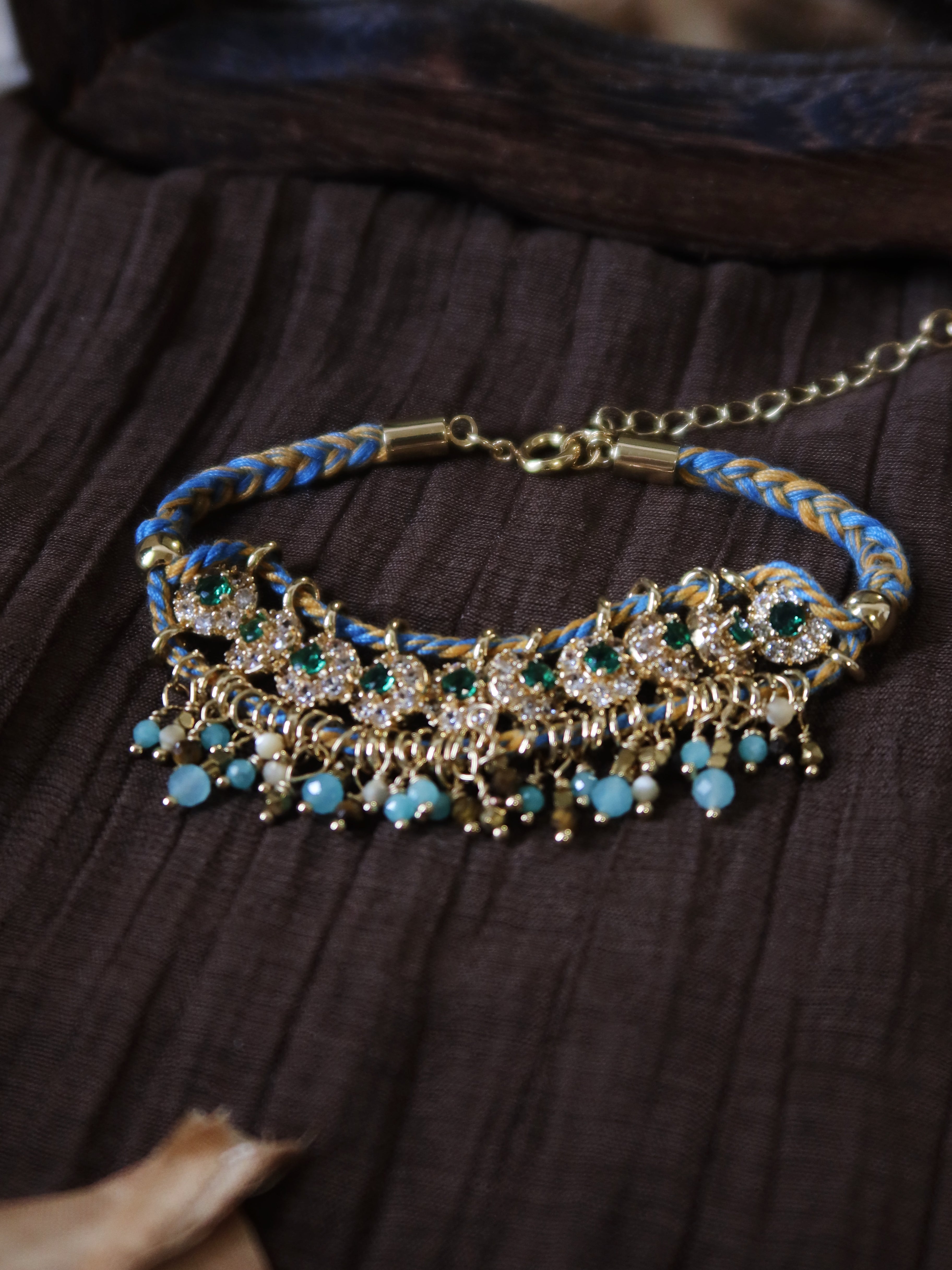 The Peacock's Muse antique green hand-woven bracelet, showcasing a structured row of green glass beads, natural tiger's eye, and micro-paved green zircons on a dark wrinkled textured fabric.
