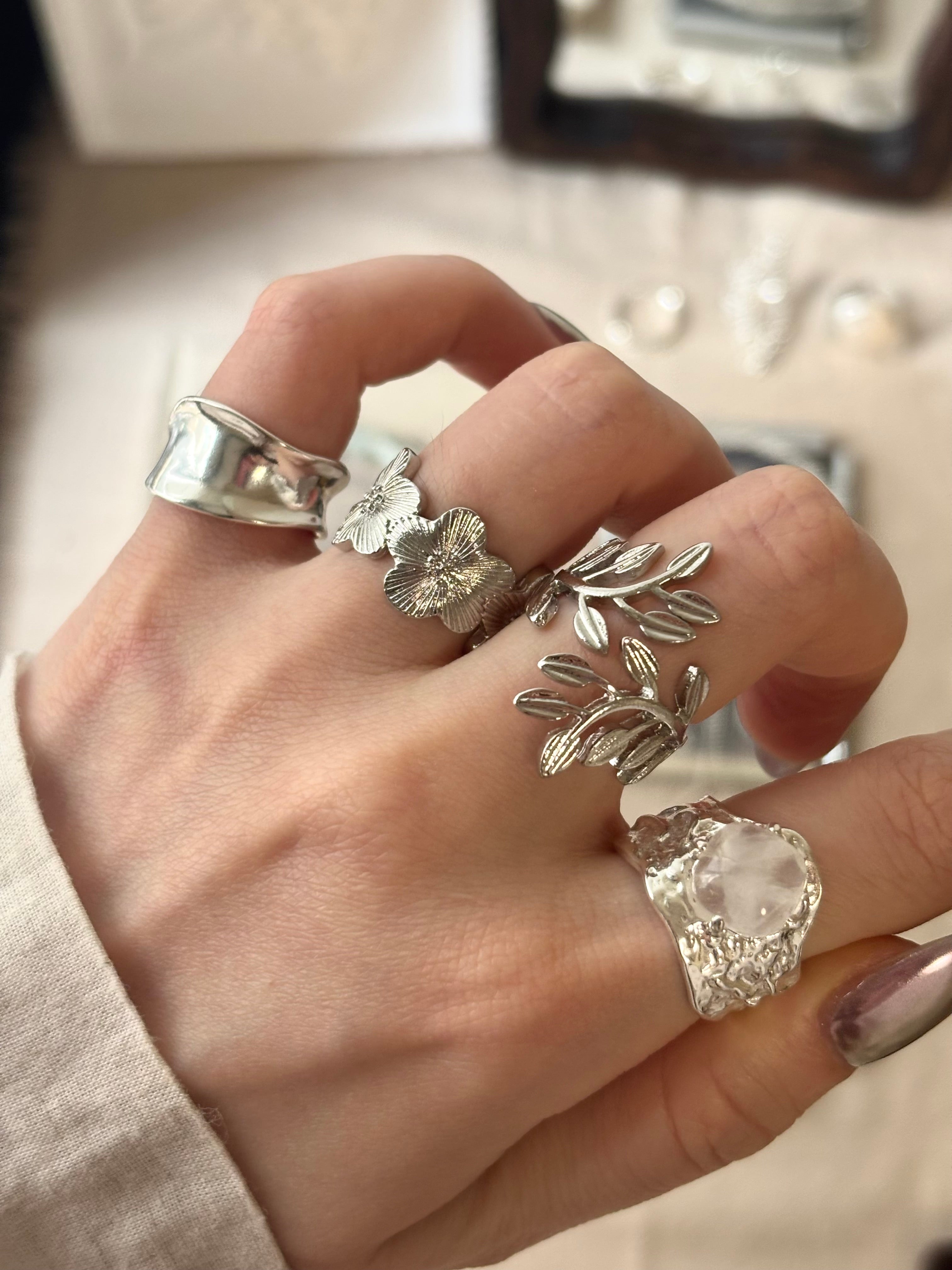 A hand wearing multiple silver rings, including a wide polished band, a flower-shaped ring, a leafy branch ring, and a textured ring with a clear crystal.
